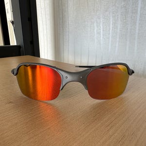 Pu&ograve; includere: Un paio di occhiali da sole con montatura grigio scuro e lenti a specchio rosso-arancio vivaci. Gli occhiali da sole sono appoggiati su una superficie di legno marrone chiaro, mostrando il loro design elegante e la loro qualit&agrave; riflettente.