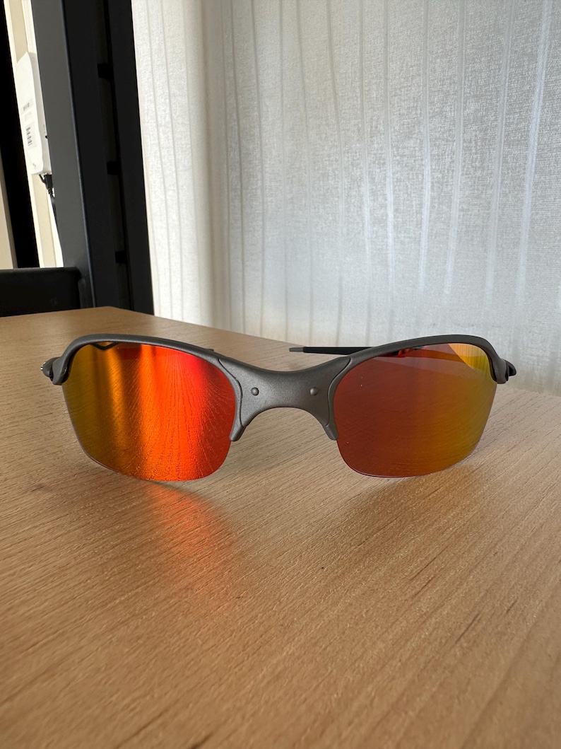 Pu&ograve; includere: Un paio di occhiali da sole con montatura grigio scuro e lenti a specchio rosso-arancio vivaci. Gli occhiali da sole sono appoggiati su una superficie di legno marrone chiaro, mostrando il loro design elegante e la loro qualit&agrave; riflettente.