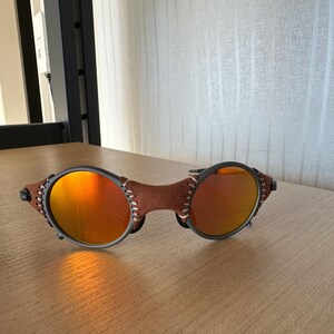 May include: Sunglasses with round orange lenses and a brown leather bridge. The frames are dark gray with visible stitching on the leather. The sunglasses are resting on a wooden surface.