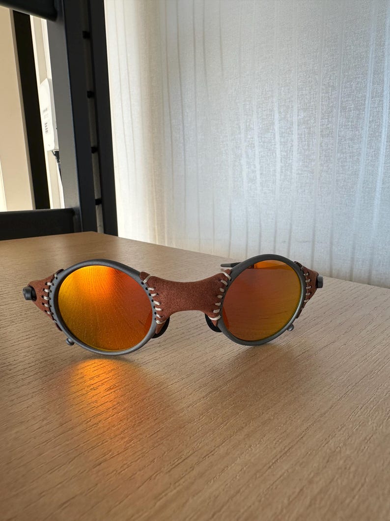 May include: Sunglasses with round orange lenses and a brown leather bridge. The frames are dark gray with visible stitching on the leather. The sunglasses are resting on a wooden surface.