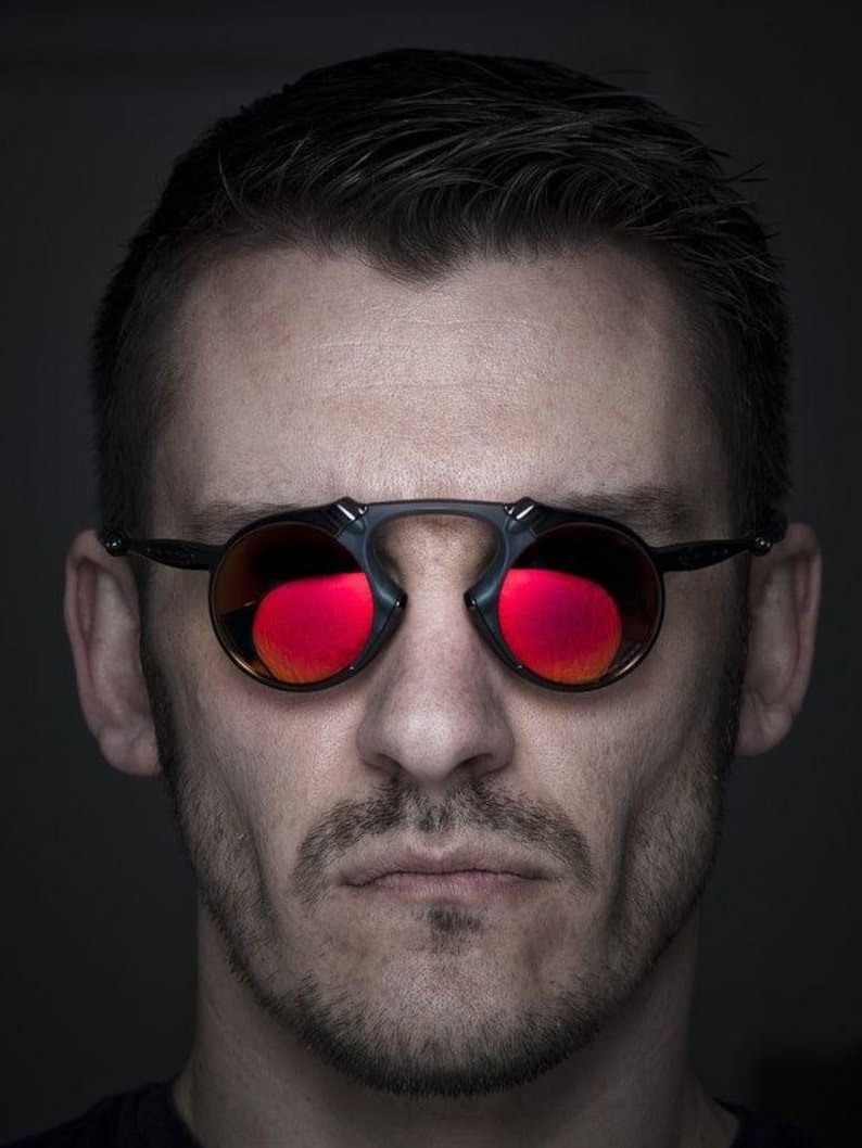Pu&ograve; includere: Primo piano di una persona con occhiali da sole rotondi con lenti rosse. Gli occhiali hanno una montatura nera e un design a ponte unico. L'immagine &egrave; un ritratto con sfondo scuro.