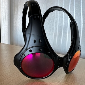 May include: Black sunglasses with a unique design, featuring a wraparound frame and red-tinted lenses. The sunglasses have a futuristic aesthetic with visible rivets and a sturdy build, suitable for outdoor activities.