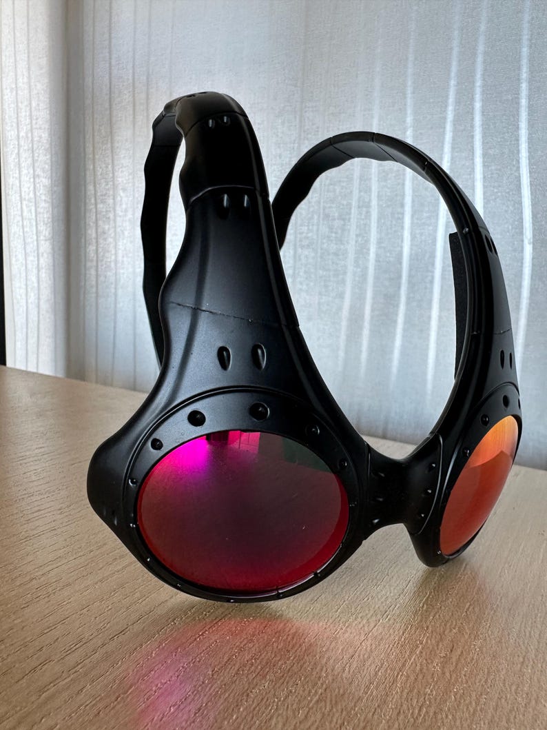 May include: Black sunglasses with a unique design, featuring a wraparound frame and red-tinted lenses. The sunglasses have a futuristic aesthetic with visible rivets and a sturdy build, suitable for outdoor activities.