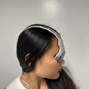 May include: Silver-coloured eyewear with a curved, futuristic design. The glasses have dark lenses and a metallic frame that curves over the forehead. The eyewear is worn by a person with long, dark hair.