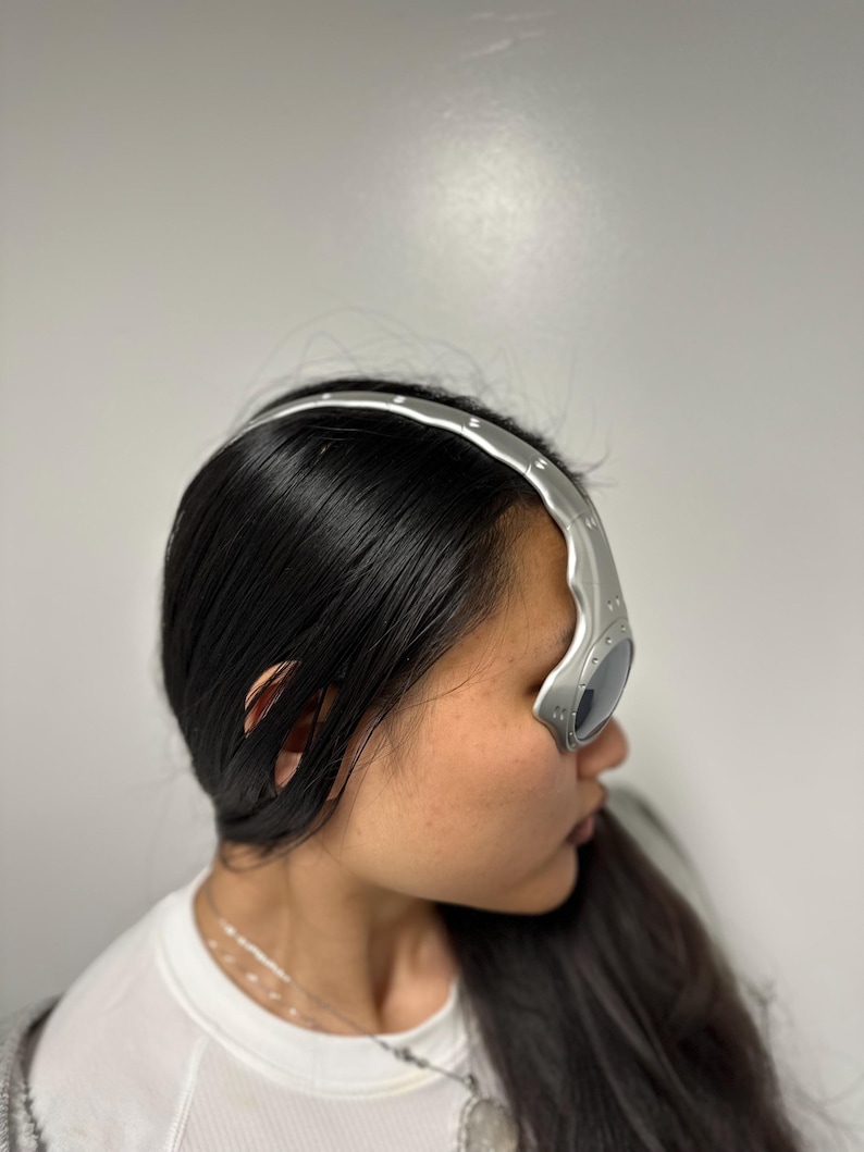 May include: Silver-coloured eyewear with a curved, futuristic design. The glasses have dark lenses and a metallic frame that curves over the forehead. The eyewear is worn by a person with long, dark hair.