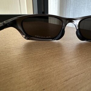 May include: Dark grey sunglasses with polarised lenses. The lenses are a dark brown colour. The frame is a dark grey colour. The word "POLARIZED" is printed on the lens.