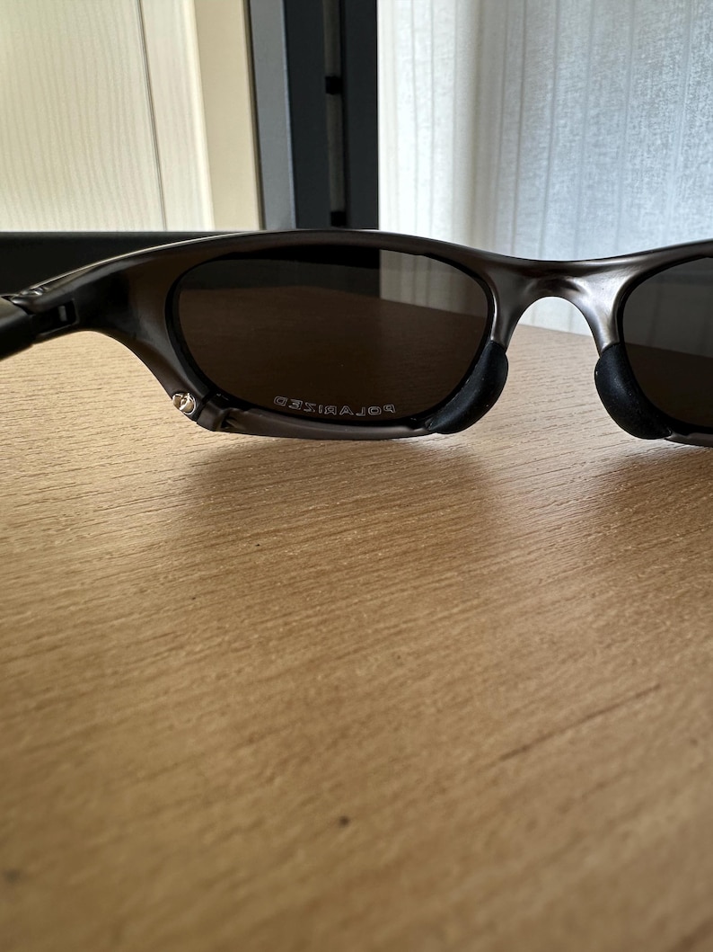 May include: Dark grey sunglasses with polarised lenses. The lenses are a dark brown colour. The frame is a dark grey colour. The word "POLARIZED" is printed on the lens.