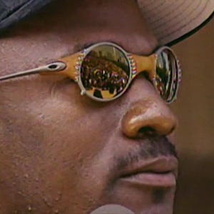 May include: Close-up of a person wearing gold-framed sunglasses with reflective lenses. The sunglasses have a unique design with a light brown frame and silver arms. The lenses reflect a crowd of people.