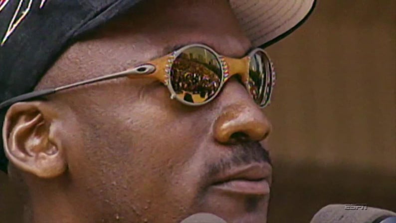 May include: Close-up of a person wearing gold-framed sunglasses with reflective lenses. The sunglasses have a unique design with a light brown frame and silver arms. The lenses reflect a crowd of people.