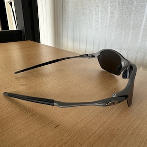Pode incluir: &Oacute;culos de sol cinza escuro com lentes pretas e hastes finas. Os &oacute;culos de sol est&atilde;o sobre uma superf&iacute;cie de madeira marrom clara. A arma&ccedil;&atilde;o tem um design elegante e moderno.