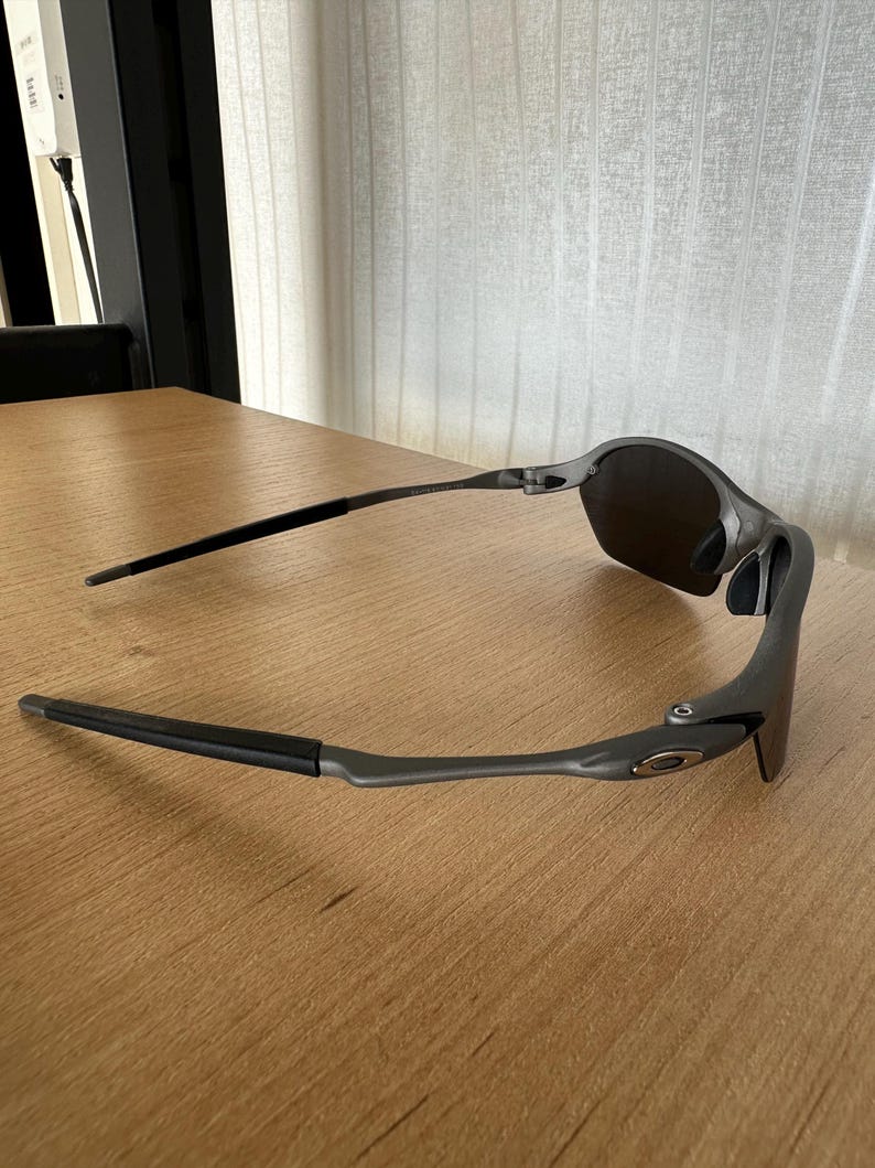 Pode incluir: &Oacute;culos de sol cinza escuro com lentes pretas e hastes finas. Os &oacute;culos de sol est&atilde;o sobre uma superf&iacute;cie de madeira marrom clara. A arma&ccedil;&atilde;o tem um design elegante e moderno.