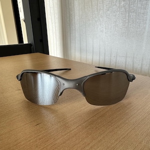 Pode incluir: Um par de &oacute;culos de sol cinzentos com lentes refletoras sobre uma superf&iacute;cie de madeira. Os &oacute;culos t&ecirc;m uma arma&ccedil;&atilde;o curva e lentes escuras, oferecendo um design elegante e moderno. A imagem est&aacute; bem iluminada.
