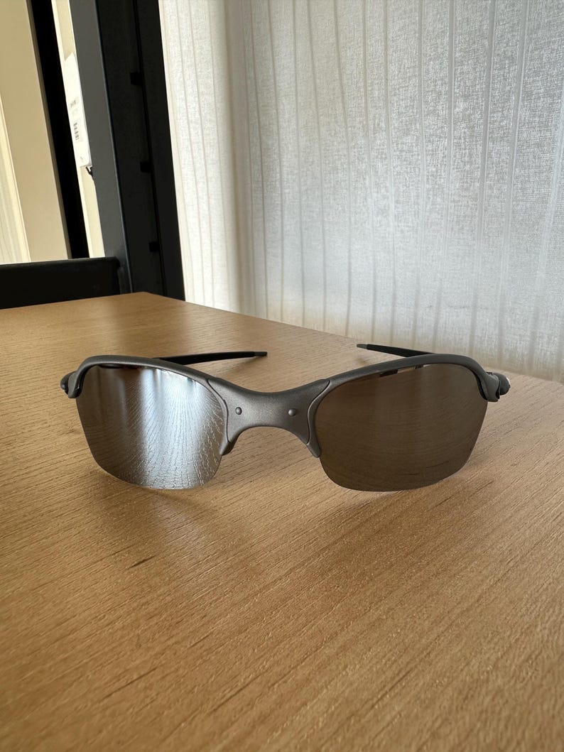 Pode incluir: Um par de &oacute;culos de sol cinzentos com lentes refletoras sobre uma superf&iacute;cie de madeira. Os &oacute;culos t&ecirc;m uma arma&ccedil;&atilde;o curva e lentes escuras, oferecendo um design elegante e moderno. A imagem est&aacute; bem iluminada.