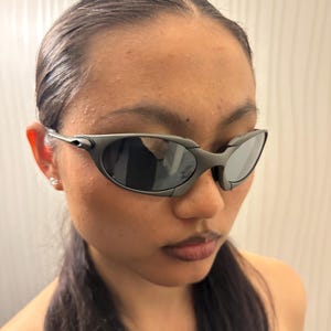 May include: Dark gray sunglasses with a sleek, wraparound design. The lenses are dark, and the frames have a matte finish. The sunglasses have a sporty, futuristic aesthetic.