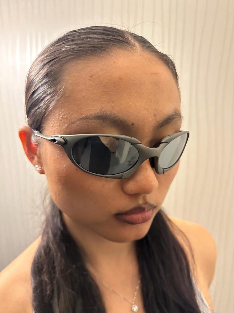 May include: Dark gray sunglasses with a sleek, wraparound design. The lenses are dark, and the frames have a matte finish. The sunglasses have a sporty, futuristic aesthetic.