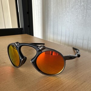 Pu&ograve; includere: Occhiali da sole neri e grigi con lenti arancioni. Gli occhiali hanno un design della montatura curvo unico e sono appoggiati su una superficie di legno. Le lenti riflettono un colore arancione brillante.