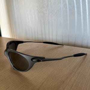 May include: Black sunglasses with a curved frame and dark lenses. The sunglasses are resting on a wooden surface. The arms of the sunglasses have a thin, sleek design.