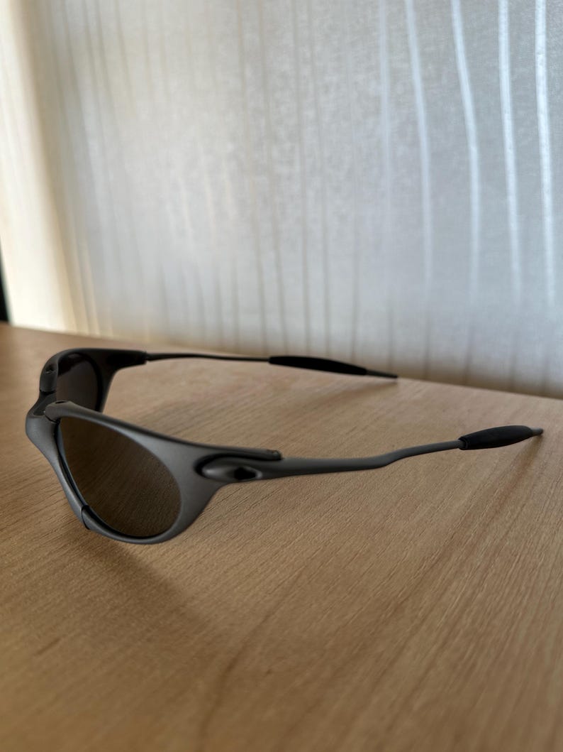 May include: Black sunglasses with a curved frame and dark lenses. The sunglasses are resting on a wooden surface. The arms of the sunglasses have a thin, sleek design.