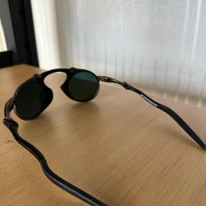 Pu&ograve; includere: Occhiali da sole neri con lenti rotonde e montatura curva. Le lenti sono di colore verde scuro. Le aste degli occhiali sono sottili e si estendono all'indietro dalla montatura.