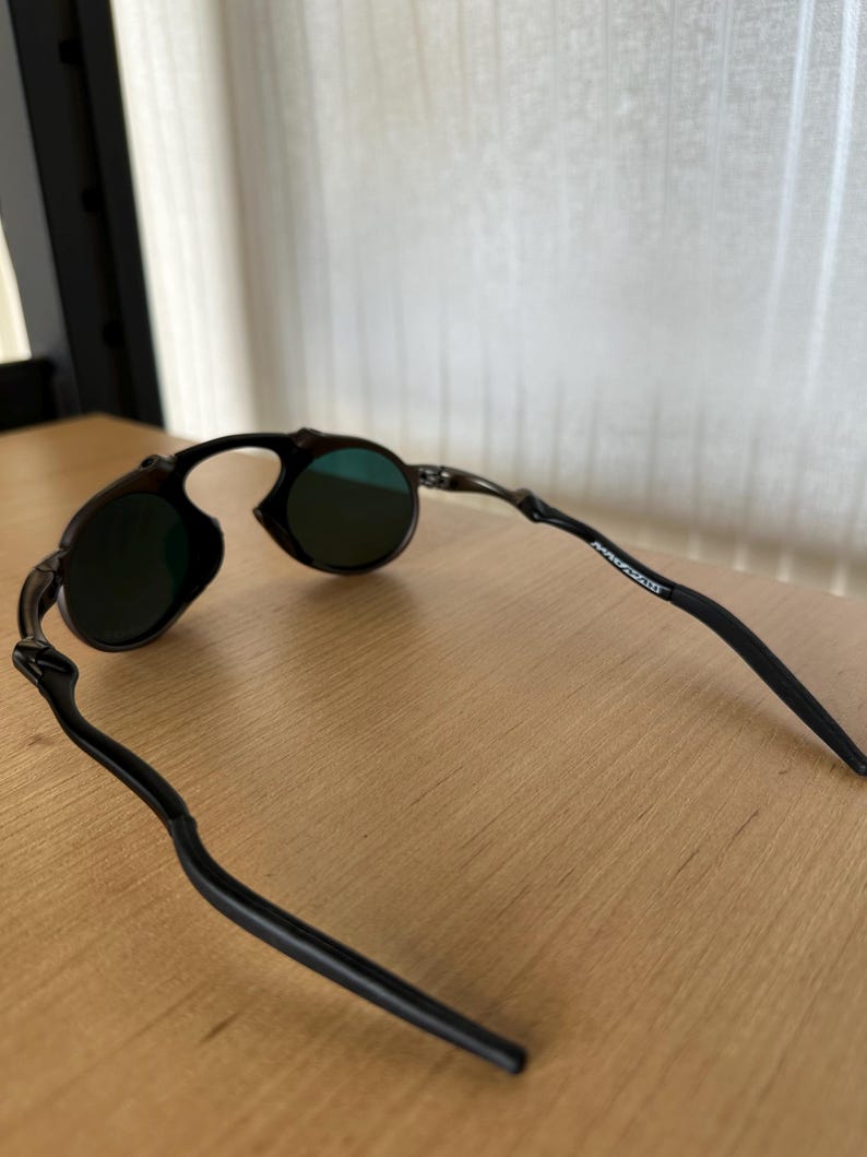 Pu&ograve; includere: Occhiali da sole neri con lenti rotonde e montatura curva. Le lenti sono di colore verde scuro. Le aste degli occhiali sono sottili e si estendono all'indietro dalla montatura.
