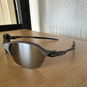 Pode incluir: &Oacute;culos de sol cinzentos com design de meia arma&ccedil;&atilde;o. As lentes s&atilde;o de cor prateada refletora e as arma&ccedil;&otilde;es s&atilde;o cinza fosco. O log&oacute;tipo da Oakley &eacute; vis&iacute;vel nas hastes. Os &oacute;culos de sol est&atilde;o sobre uma superf&iacute;cie de madeira.