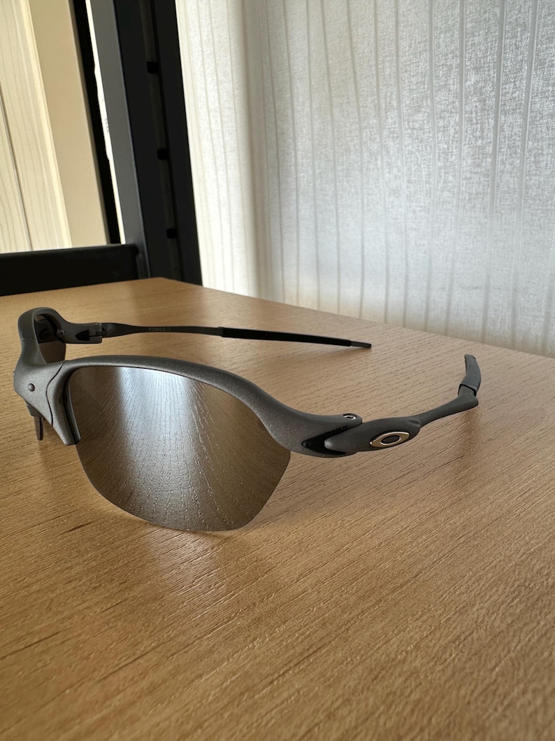Pode incluir: &Oacute;culos de sol cinzentos com design de meia arma&ccedil;&atilde;o. As lentes s&atilde;o de cor prateada refletora e as arma&ccedil;&otilde;es s&atilde;o cinza fosco. O log&oacute;tipo da Oakley &eacute; vis&iacute;vel nas hastes. Os &oacute;culos de sol est&atilde;o sobre uma superf&iacute;cie de madeira.