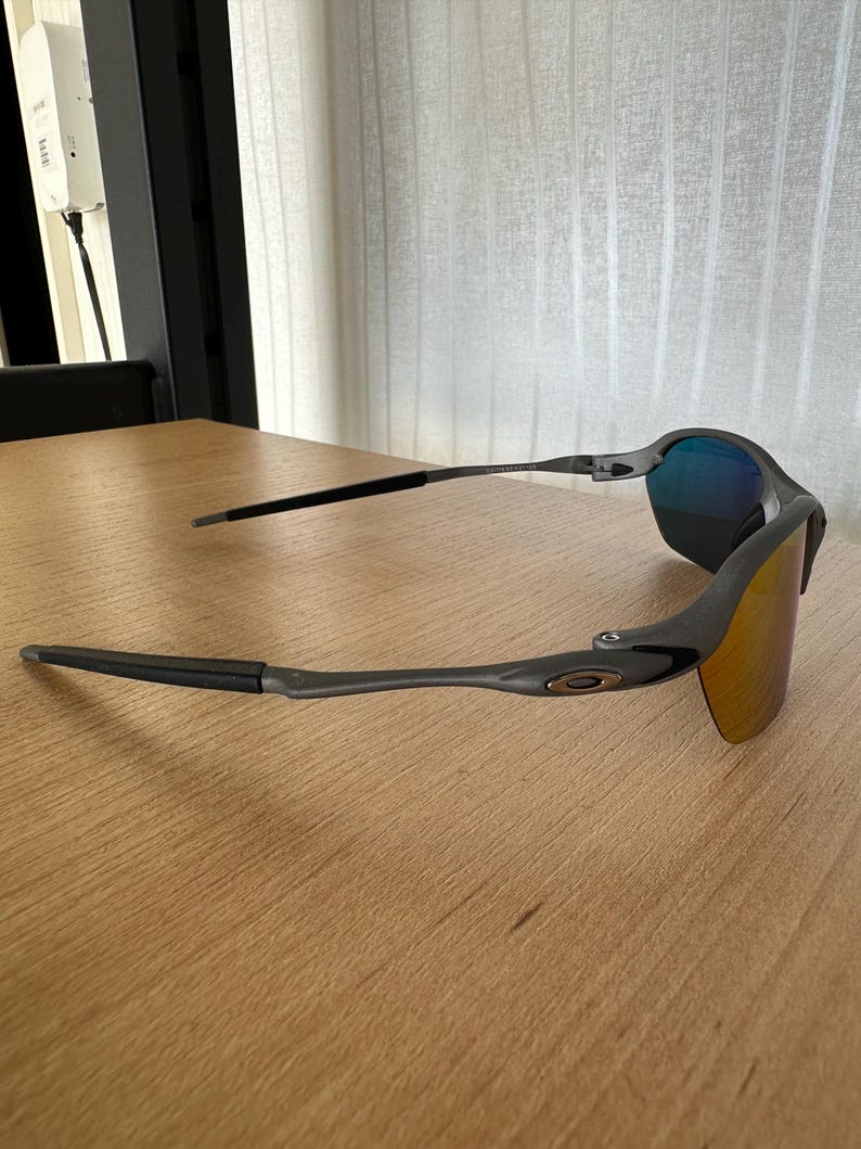 Pu&ograve; includere: Occhiali da sole grigi e neri con lenti a specchio. Gli occhiali hanno un design elegante e moderno con una montatura curva e aste scure. Le lenti riflettono sfumature blu e arancioni.