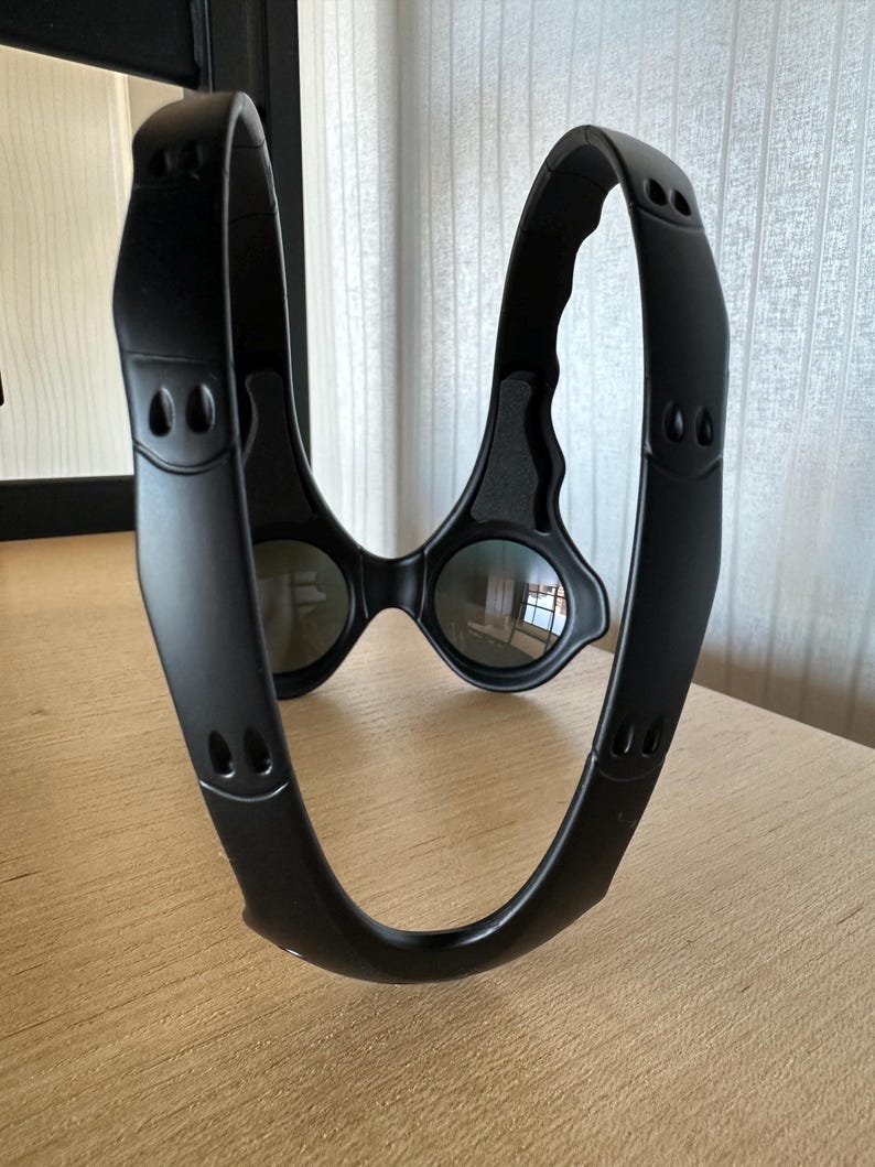 May include: Black sports eyewear with a unique, wraparound design. The glasses feature dark lenses and a thick, curved frame with small, decorative cutouts. The frame appears to be made of a matte material.