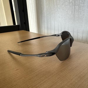 Pode incluir: &Oacute;culos de sol cinzentos com lentes escuras. Os &oacute;culos t&ecirc;m um design elegante e moderno com uma arma&ccedil;&atilde;o e hastes curvas. As lentes s&atilde;o de uma cor escura e refletora, e a arma&ccedil;&atilde;o &eacute; cinza fosco.