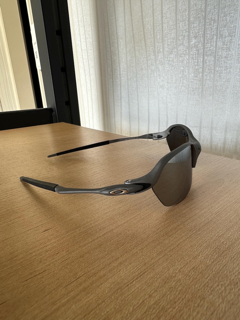Pode incluir: &Oacute;culos de sol cinzentos com lentes escuras. Os &oacute;culos t&ecirc;m um design elegante e moderno com uma arma&ccedil;&atilde;o e hastes curvas. As lentes s&atilde;o de uma cor escura e refletora, e a arma&ccedil;&atilde;o &eacute; cinza fosco.