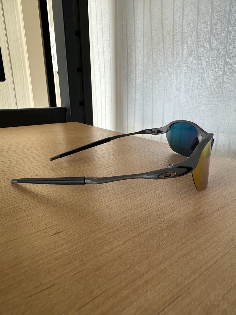 Pu&ograve; includere: Occhiali da sole neri dal design elegante e moderno. Le lenti hanno una finitura a specchio con sfumature blu e oro. Le montature sono grigio scuro e presentano un logo discreto sulle aste. Gli occhiali sono appoggiati su una superficie di legno.