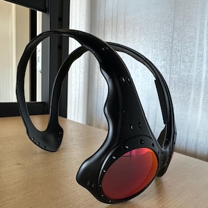 May include: Black sunglasses with red lenses. The sunglasses have a unique, wraparound design with a thick frame and visible rivets. The lenses are a vibrant red color, and the frame is matte black.