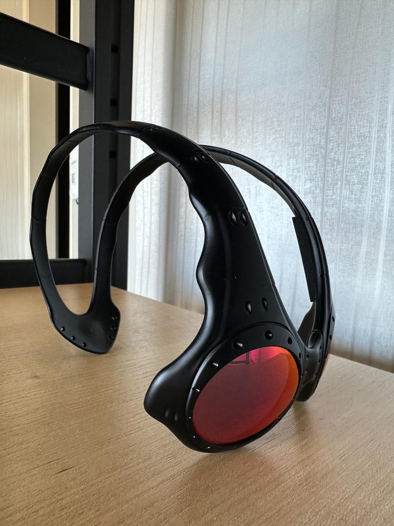 May include: Black sunglasses with red lenses. The sunglasses have a unique, wraparound design with a thick frame and visible rivets. The lenses are a vibrant red color, and the frame is matte black.