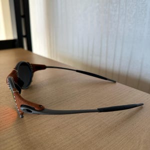 May include: Sunglasses with a dark frame and lenses. The temples are brown leather with white stitching. The lenses are round and dark, and the frame is a dark gray color. The sunglasses are resting on a wooden surface.
