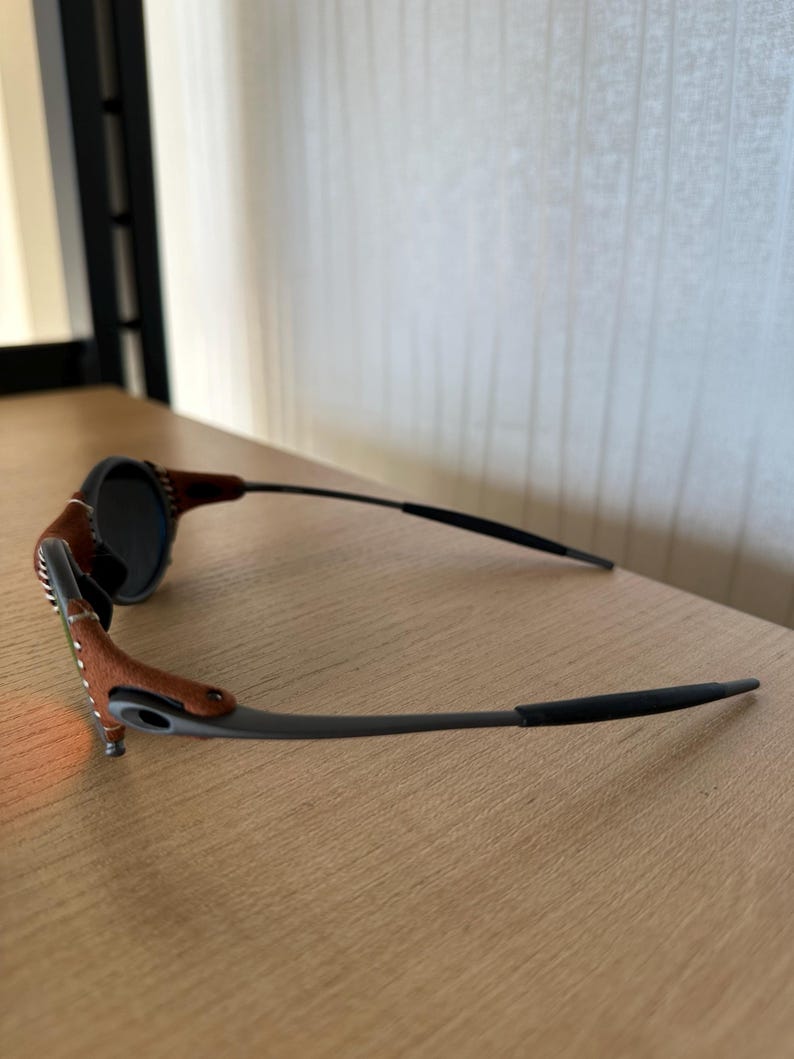 May include: Sunglasses with a dark frame and lenses. The temples are brown leather with white stitching. The lenses are round and dark, and the frame is a dark gray color. The sunglasses are resting on a wooden surface.
