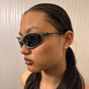 May include: Gray sunglasses with dark lenses. The sunglasses have a wraparound design with a sleek, modern aesthetic. The frame is a matte gray color, and the lenses are dark, providing a contrast. The temples are thin and curve around the ears.