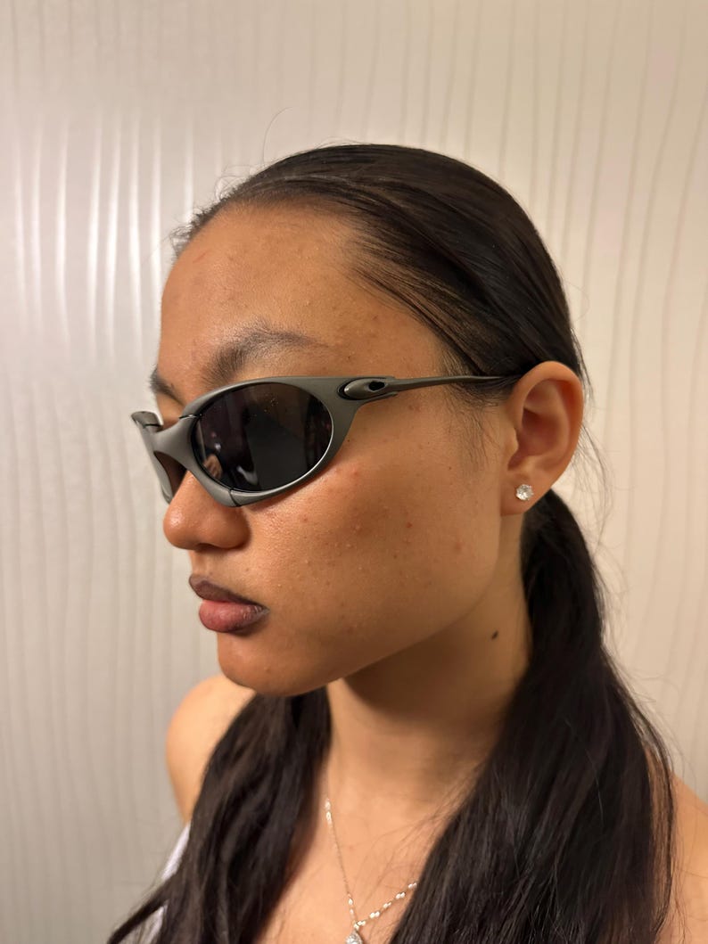 May include: Gray sunglasses with dark lenses. The sunglasses have a wraparound design with a sleek, modern aesthetic. The frame is a matte gray color, and the lenses are dark, providing a contrast. The temples are thin and curve around the ears.