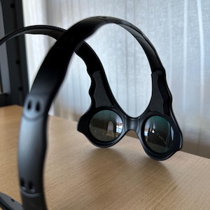 May include: Black, futuristic-style glasses with a unique frame design. The lenses are round and reflect the surrounding environment. The frame curves over the head and has a sleek, modern aesthetic. The glasses are resting on a wooden surface.