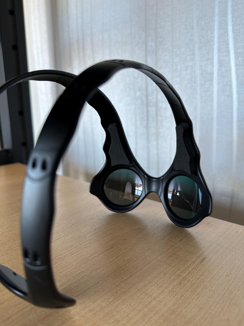 May include: Black, futuristic-style glasses with a unique frame design. The lenses are round and reflect the surrounding environment. The frame curves over the head and has a sleek, modern aesthetic. The glasses are resting on a wooden surface.
