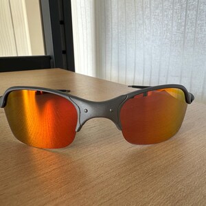 Pu&ograve; includere: Occhiali da sole grigio scuro con lenti a specchio arancione-rosse. Gli occhiali hanno un design semi-senza montatura e una montatura curva. Le lenti riflettono la luce ambientale, creando un gradiente arancione e giallo.