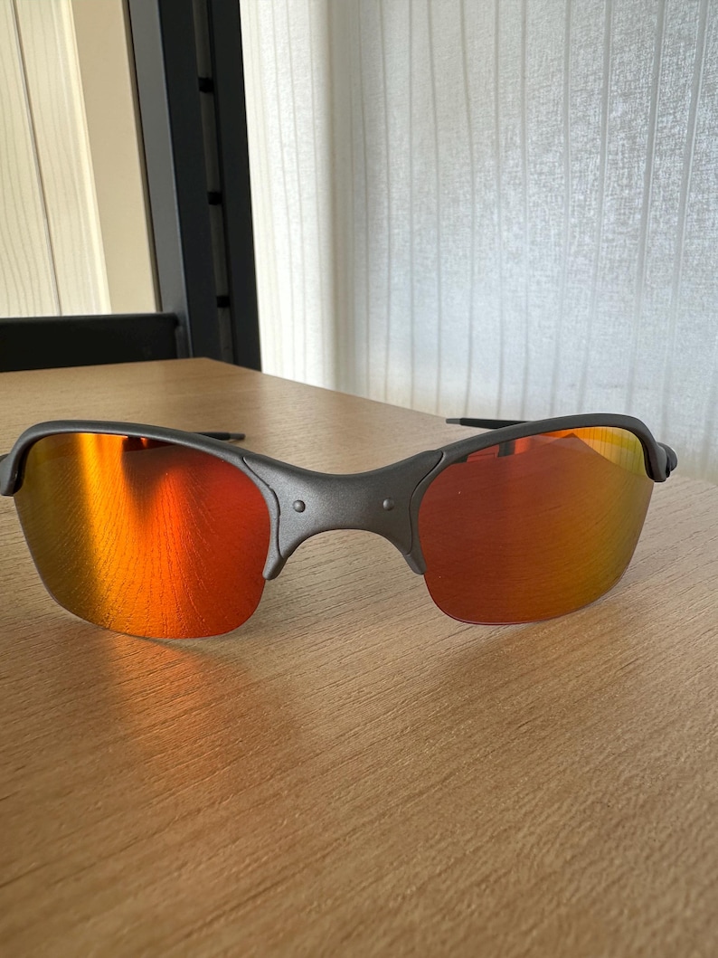 Pu&ograve; includere: Occhiali da sole grigio scuro con lenti a specchio arancione-rosse. Gli occhiali hanno un design semi-senza montatura e una montatura curva. Le lenti riflettono la luce ambientale, creando un gradiente arancione e giallo.