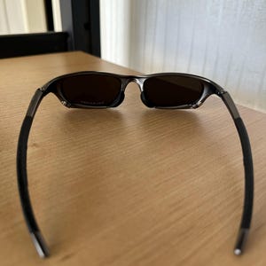May include: Dark sunglasses with tinted lenses. The frame is a dark metallic colour with black rubberised temple arms. The sunglasses are resting on a light brown wooden surface.