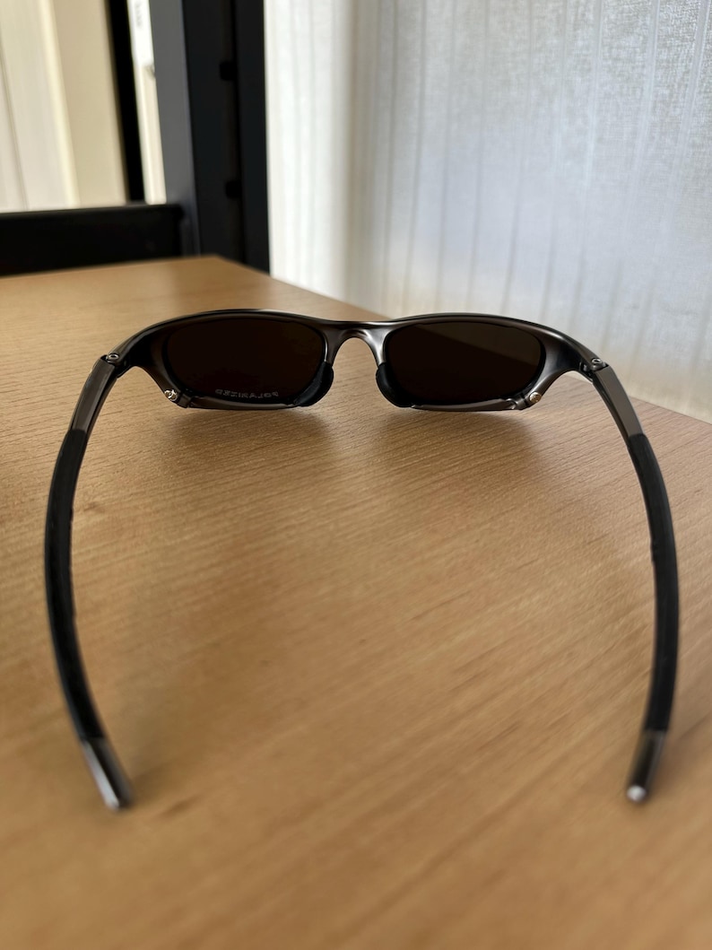 May include: Dark sunglasses with tinted lenses. The frame is a dark metallic colour with black rubberised temple arms. The sunglasses are resting on a light brown wooden surface.