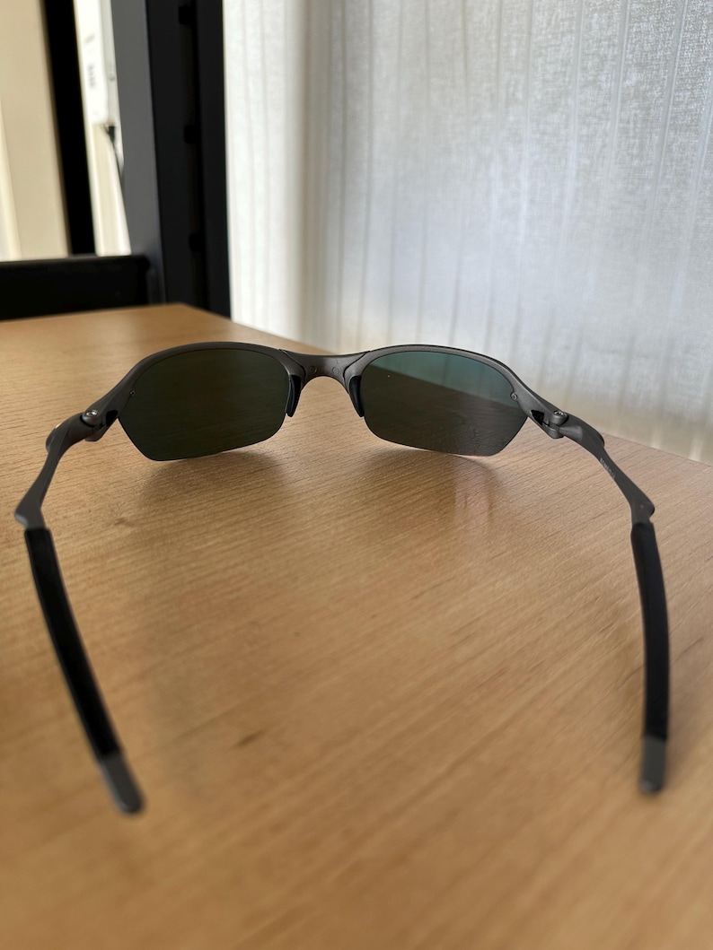 Pu&ograve; includere: Occhiali da sole grigio scuro con lenti verdi. Gli occhiali da sole hanno un design elegante e moderno con una montatura sottile e aste curve. Le lenti sono di colore verde scuro.