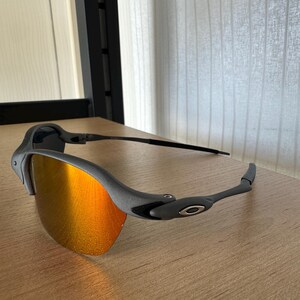 Pu&ograve; includere: Occhiali da sole grigi con lenti a specchio arancioni. Gli occhiali hanno un design elegante con una montatura e aste grigio scuro. Le lenti riflettono un colore arancione brillante e il logo Oakley &egrave; visibile sull'asta.