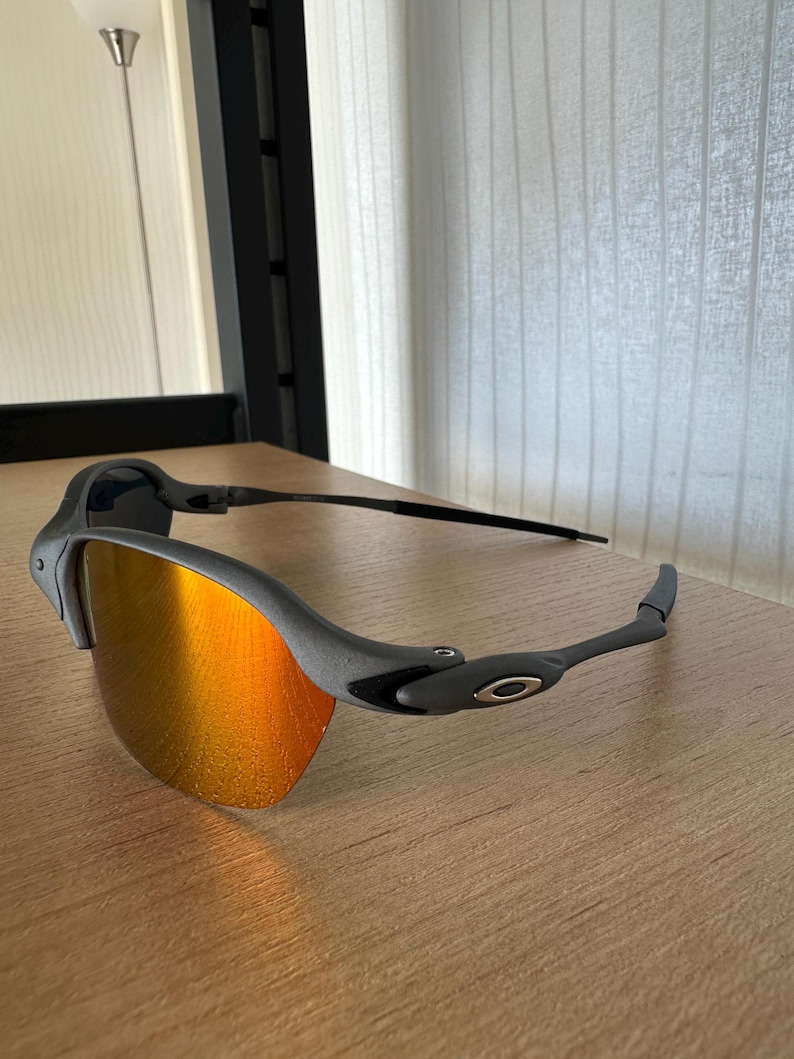 Pu&ograve; includere: Occhiali da sole grigi con lenti a specchio arancioni. Gli occhiali hanno un design elegante con una montatura e aste grigio scuro. Le lenti riflettono un colore arancione brillante e il logo Oakley &egrave; visibile sull'asta.