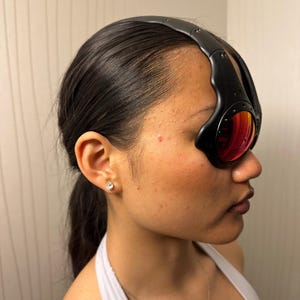 May include: Black sport sunglasses with red-tinted lenses. The sunglasses have a thick, curved black frame that wraps around the head. The lenses are oval-shaped and reflect a reddish-orange hue.