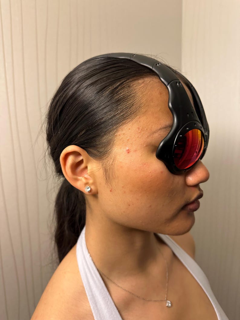 May include: Black sport sunglasses with red-tinted lenses. The sunglasses have a thick, curved black frame that wraps around the head. The lenses are oval-shaped and reflect a reddish-orange hue.