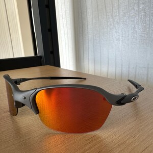 Pu&ograve; includere: Un paio di occhiali da sole grigio opaco con lenti rosso-arancio vivaci. Gli occhiali hanno un design elegante e moderno con una montatura curva e un piccolo logo sulle aste. Le lenti sono riflettenti.