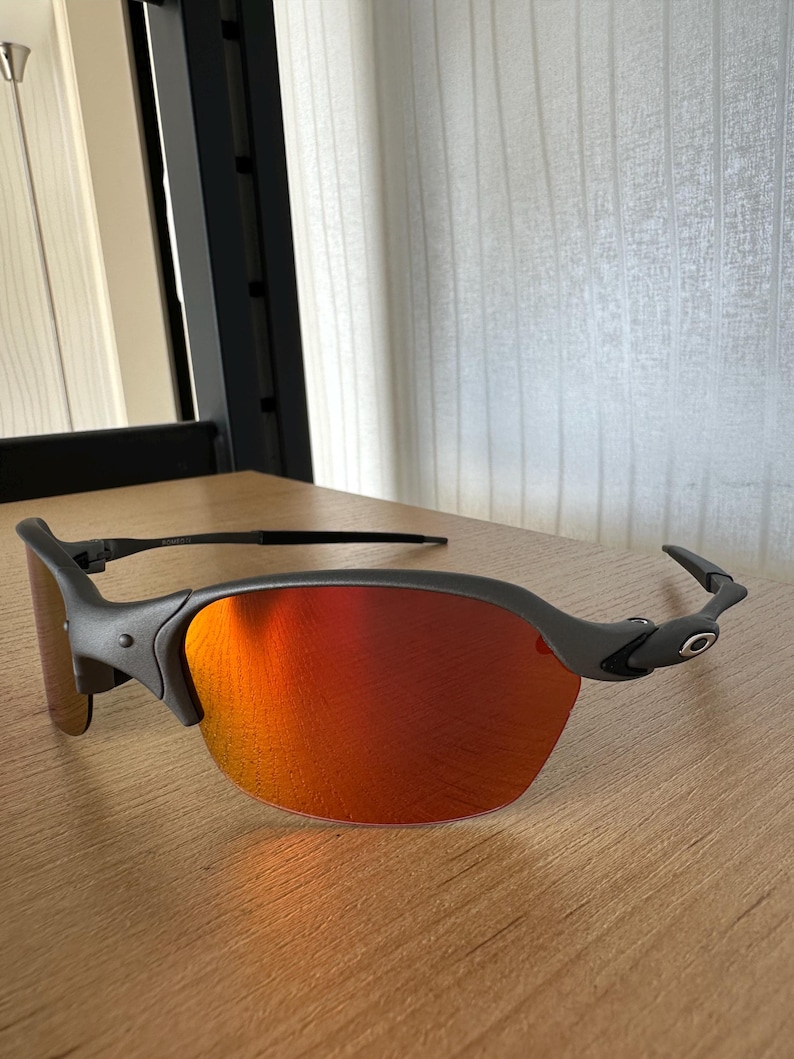 Pu&ograve; includere: Un paio di occhiali da sole grigio opaco con lenti rosso-arancio vivaci. Gli occhiali hanno un design elegante e moderno con una montatura curva e un piccolo logo sulle aste. Le lenti sono riflettenti.